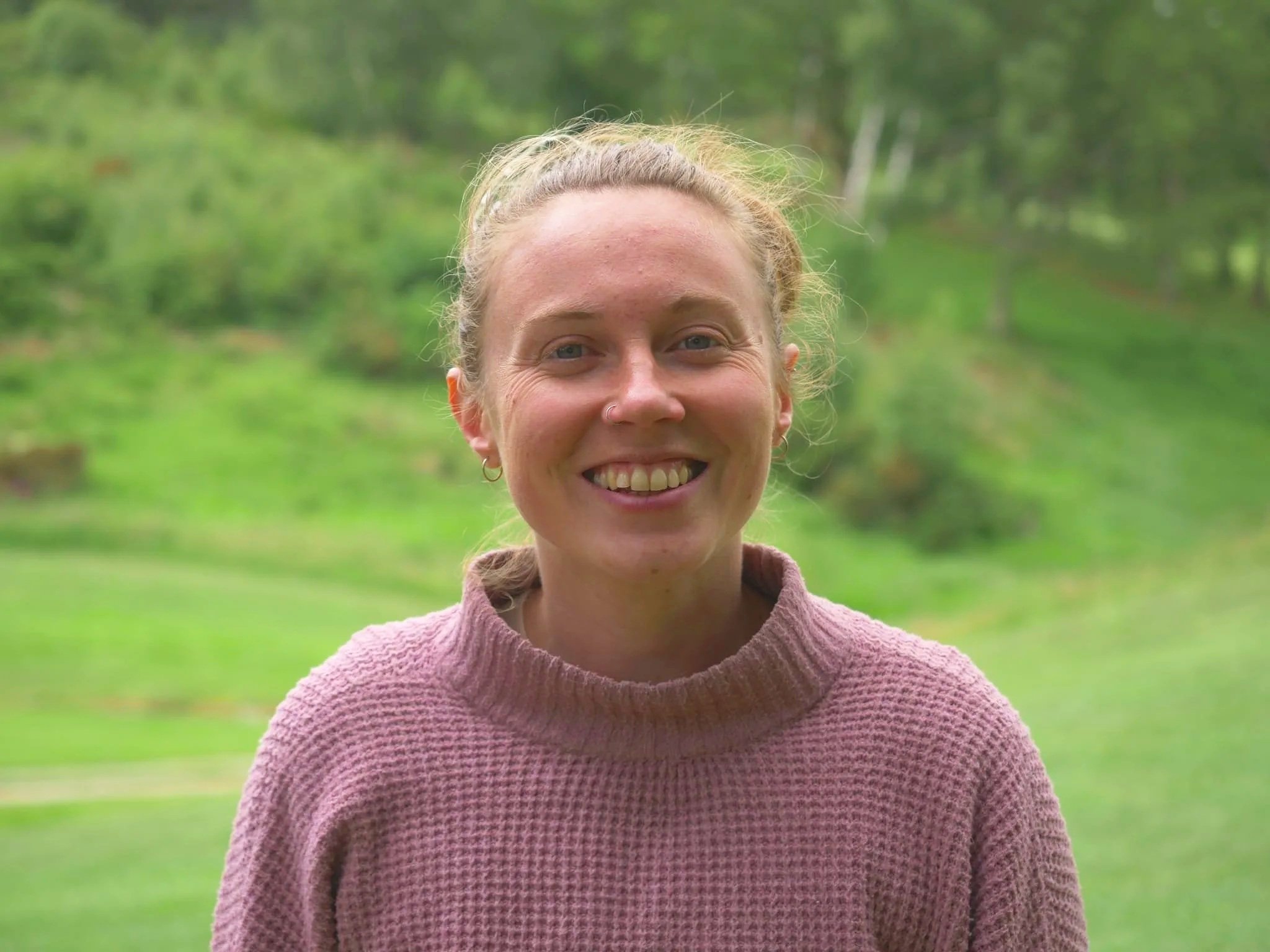 Lucy Gray - A woman with a light brown hair tied back, smiling, outdoors in a green, wooded area, wearing a pink, textured sweater.