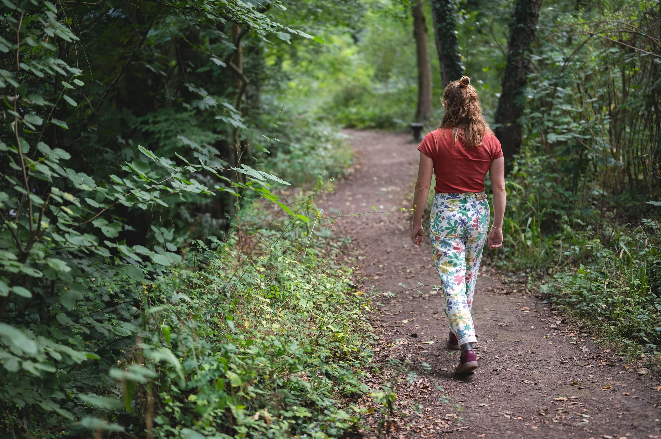 A woman with brown, curly hair tied in a bun, wearing a red T-shirt and colorful floral pants, is walking on a dirt trail through a green forest surrounded by lush trees and bushes.