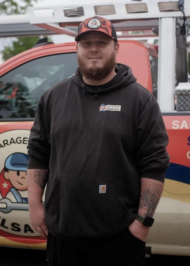 A man with a beard and tattoos wearing a black hoodie and a red and black cap, standing in front of a utility truck.