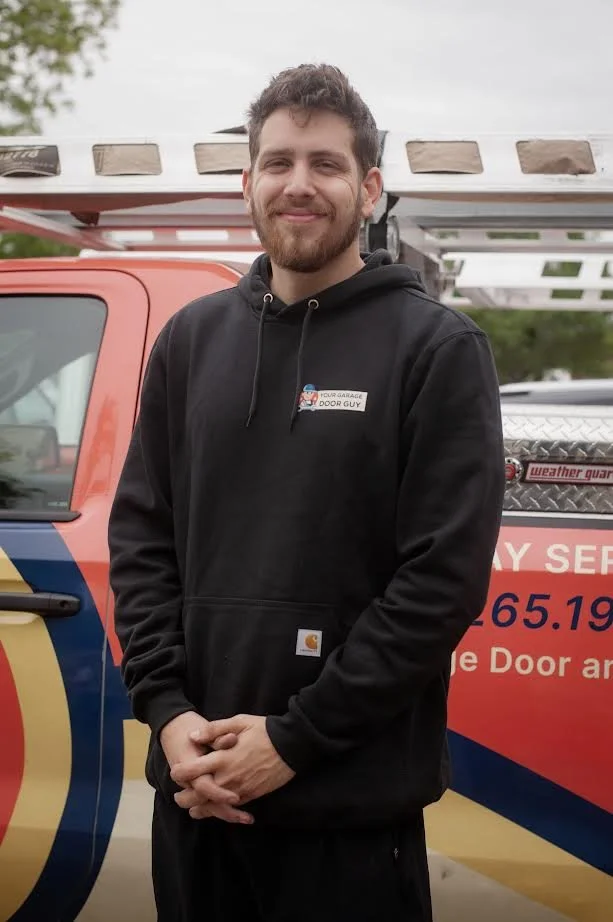 A young man with a beard and mustache wearing a black Carhartt hoodie standing in front of a red and blue service truck with ladder on top, outdoors.