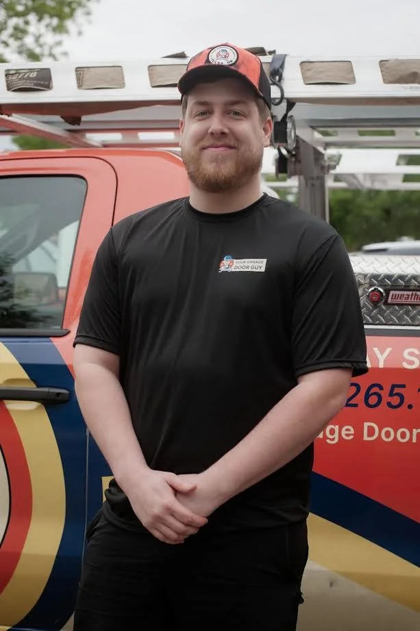 A man with a beard and wearing a black shirt and cap standing in front of a red and blue utility truck with a ladder attached to it.