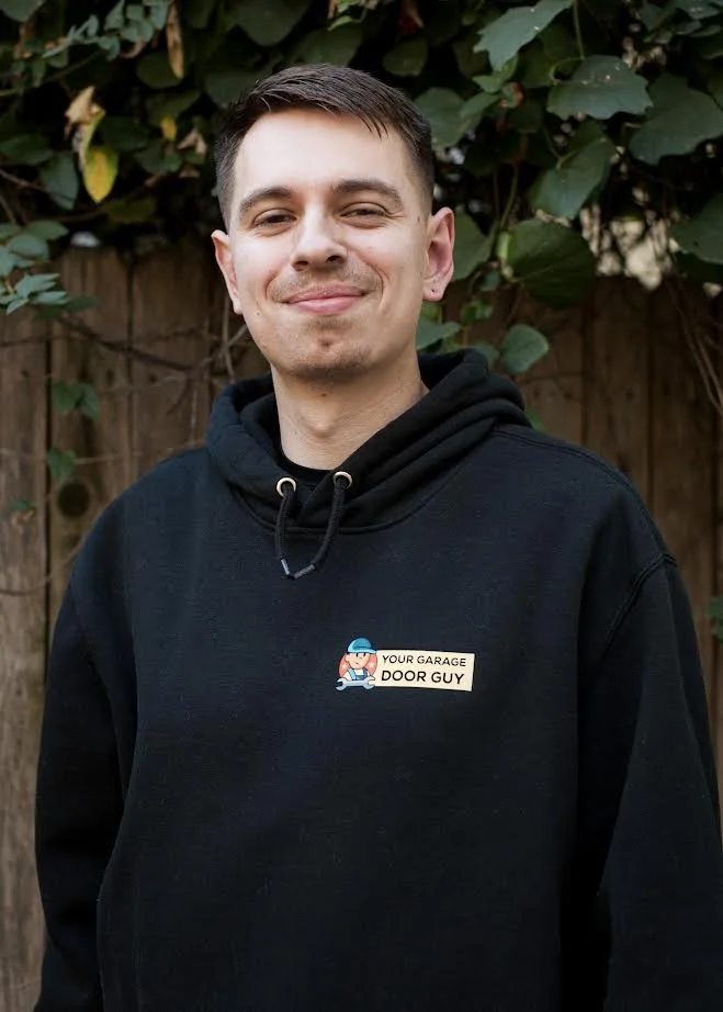 Young man smiling outdoors, wearing a black hoodie with a badge that says 'Your Garage Door Guy' in front of a wooden fence and leafy plants.