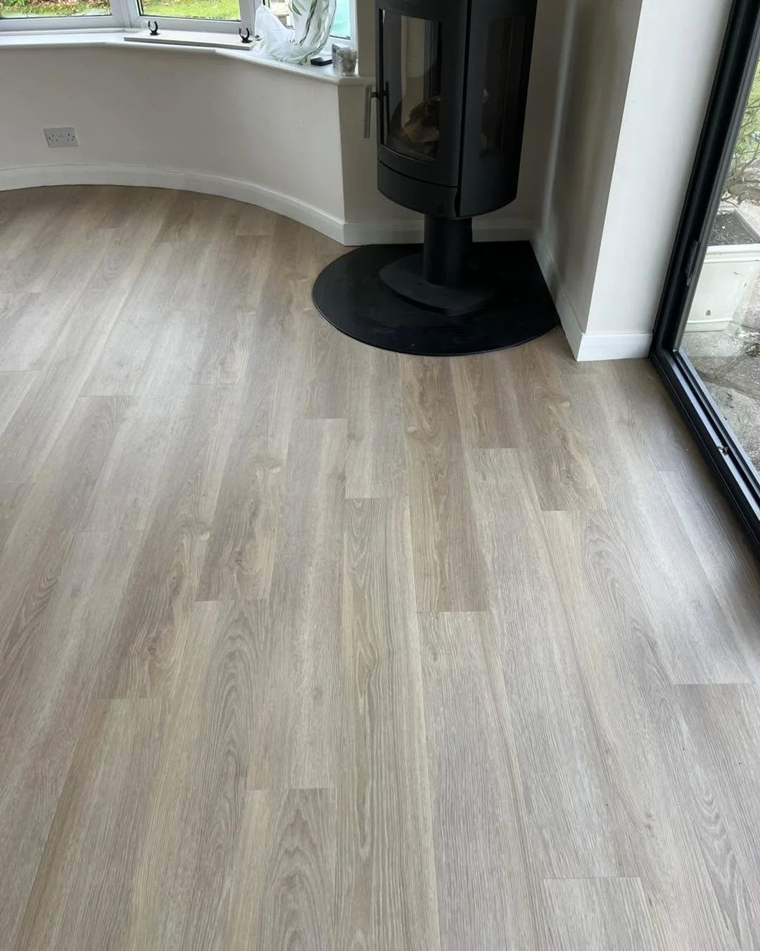 LVT flooring in a corner of a room with a black fireplace stove on a black oval mat, near a large glass door and a curved white wall with a window sill.