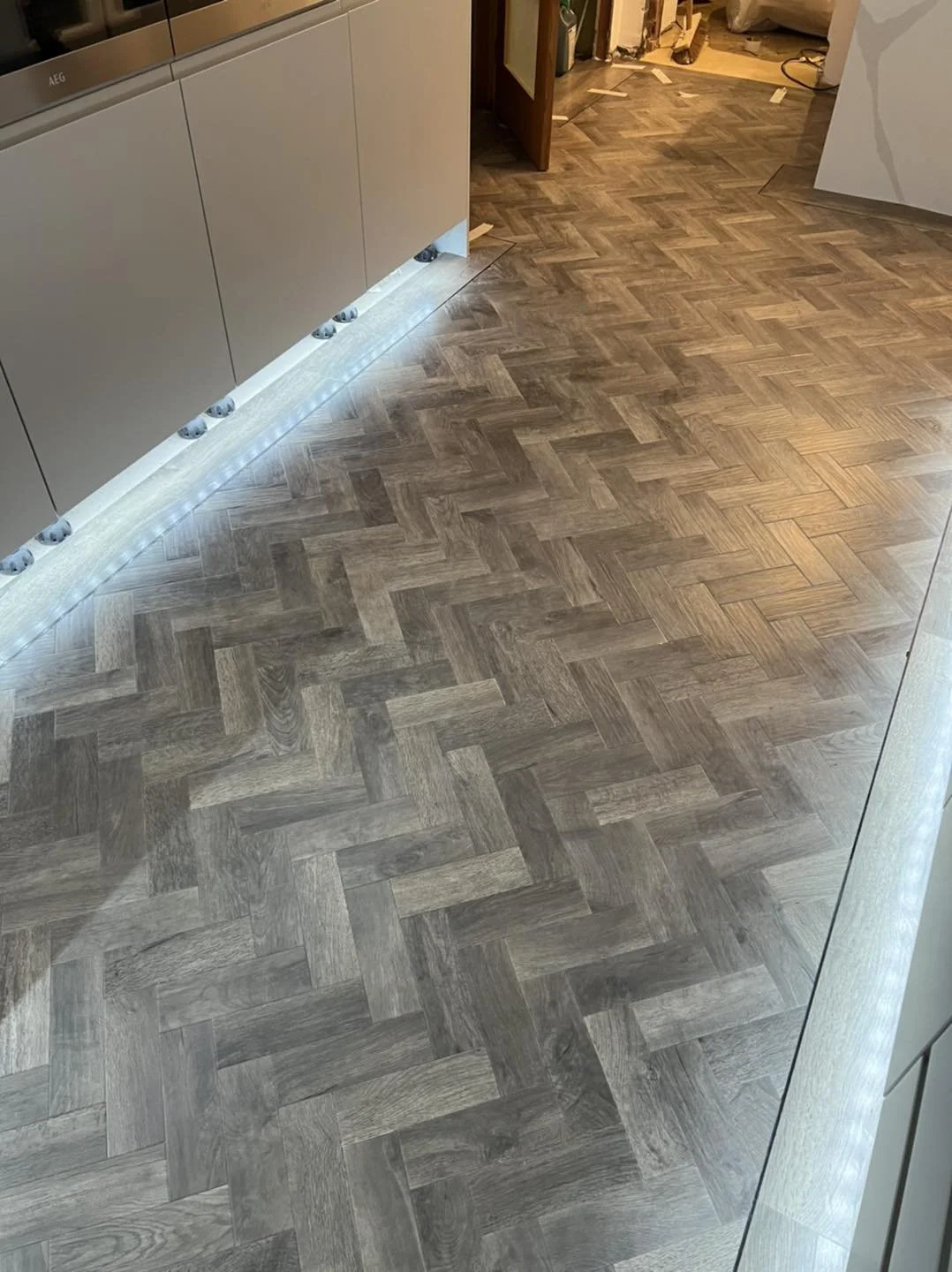 Interior of a room with a new wooden herringbone patterned floor, under-cabinet lighting reflects on the surface, and construction materials are visible in the background.