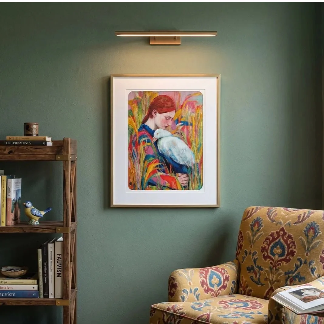 A framed colorful painting of a woman with red hair holding a white bird on her shoulder, hanging on a green wall with a modern light above it. To the left, a wooden bookshelf with books and decorative items, and a patterned armchair with a magazine on it in a cozy room.