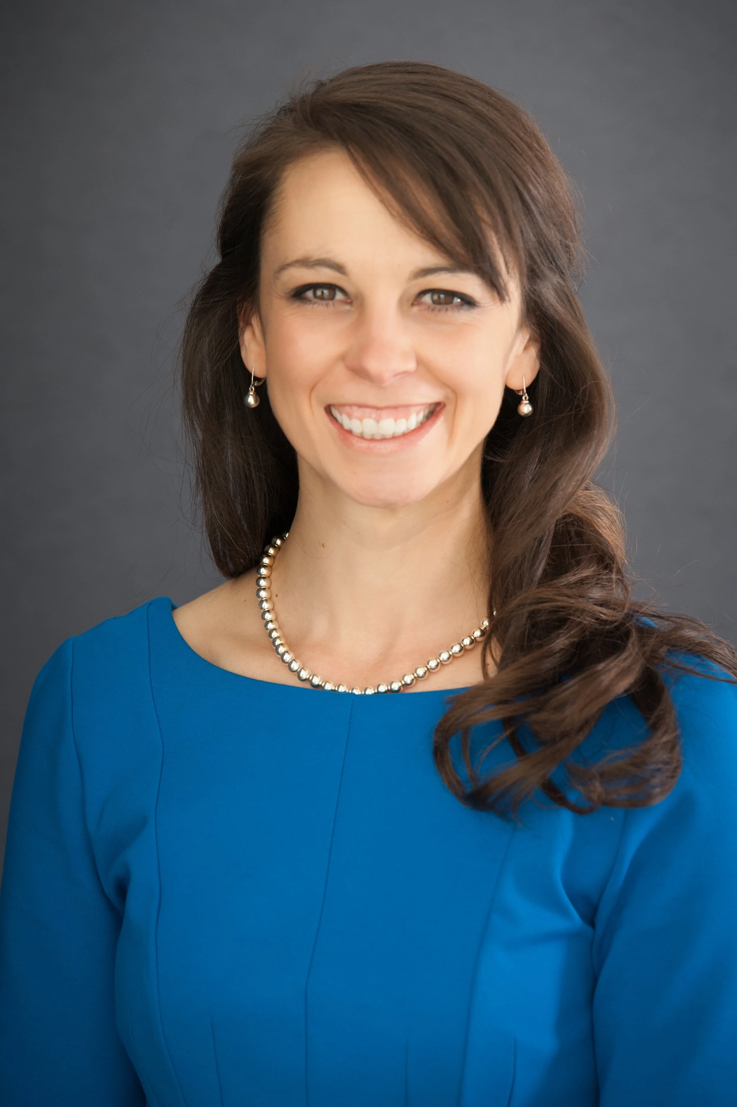 Portrait of a smiling woman with long brown hair, wearing a blue dress, pearl necklace, and pearl earrings, against a dark gray background.