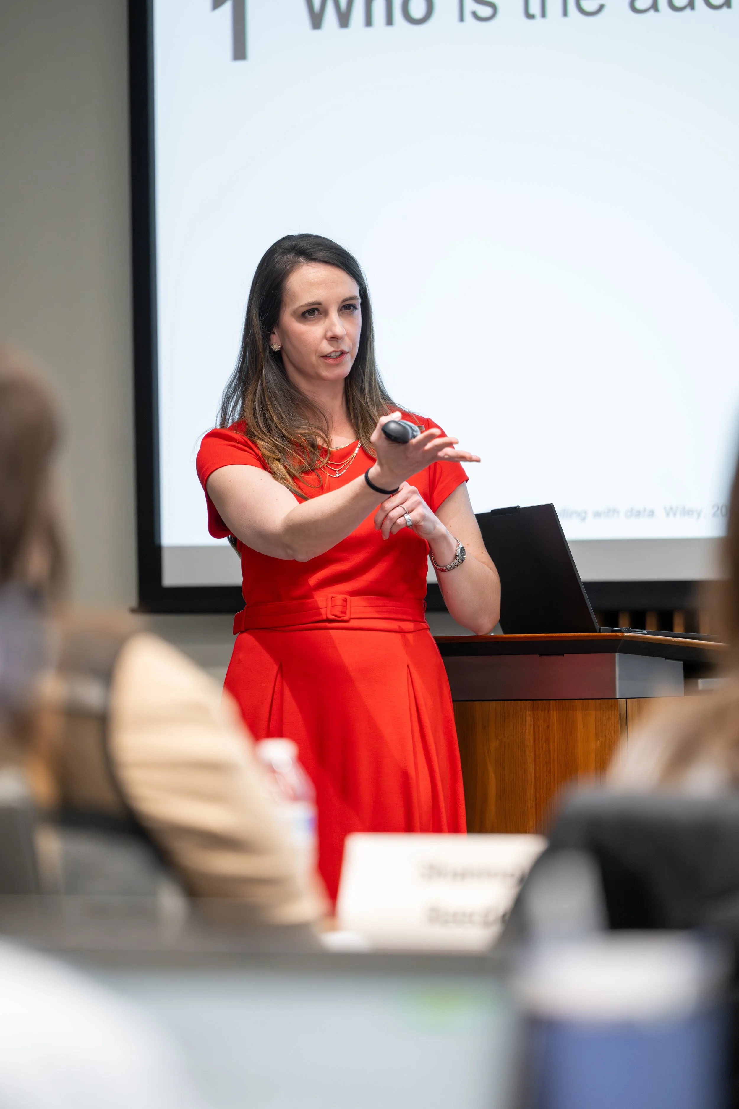 A woman in a red dress giving a presentation in a conference room with a large screen displaying a slide.
