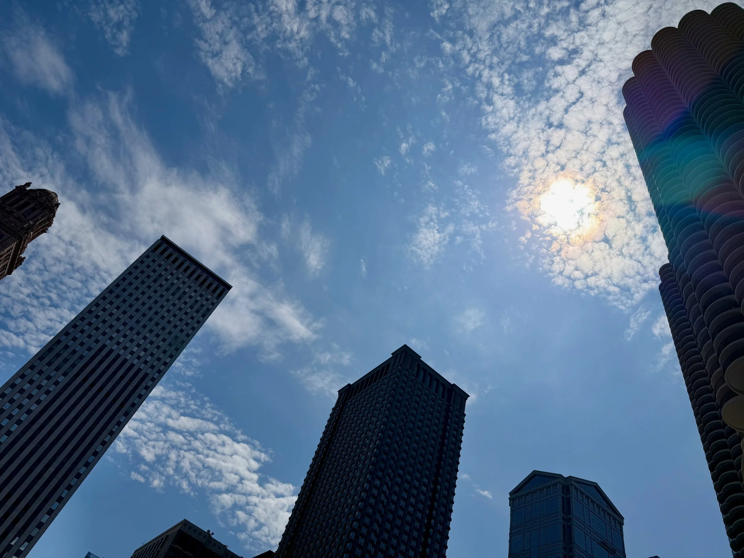View of several modern skyscrapers reaching towards a partly cloudy sky with the sun shining behind the clouds.