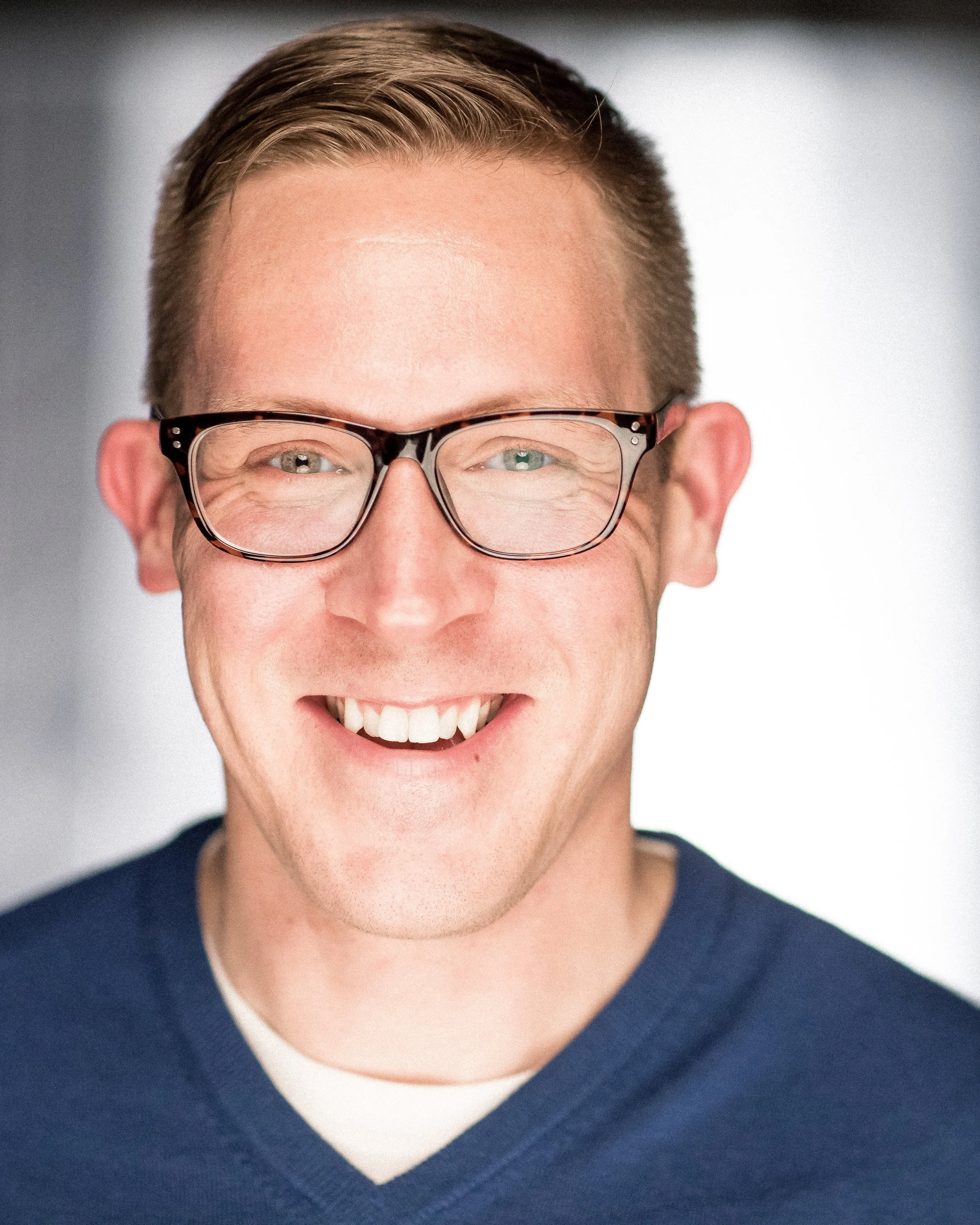 Close-up of smiling person with short hair, wearing glasses and a navy sweater over a white shirt.