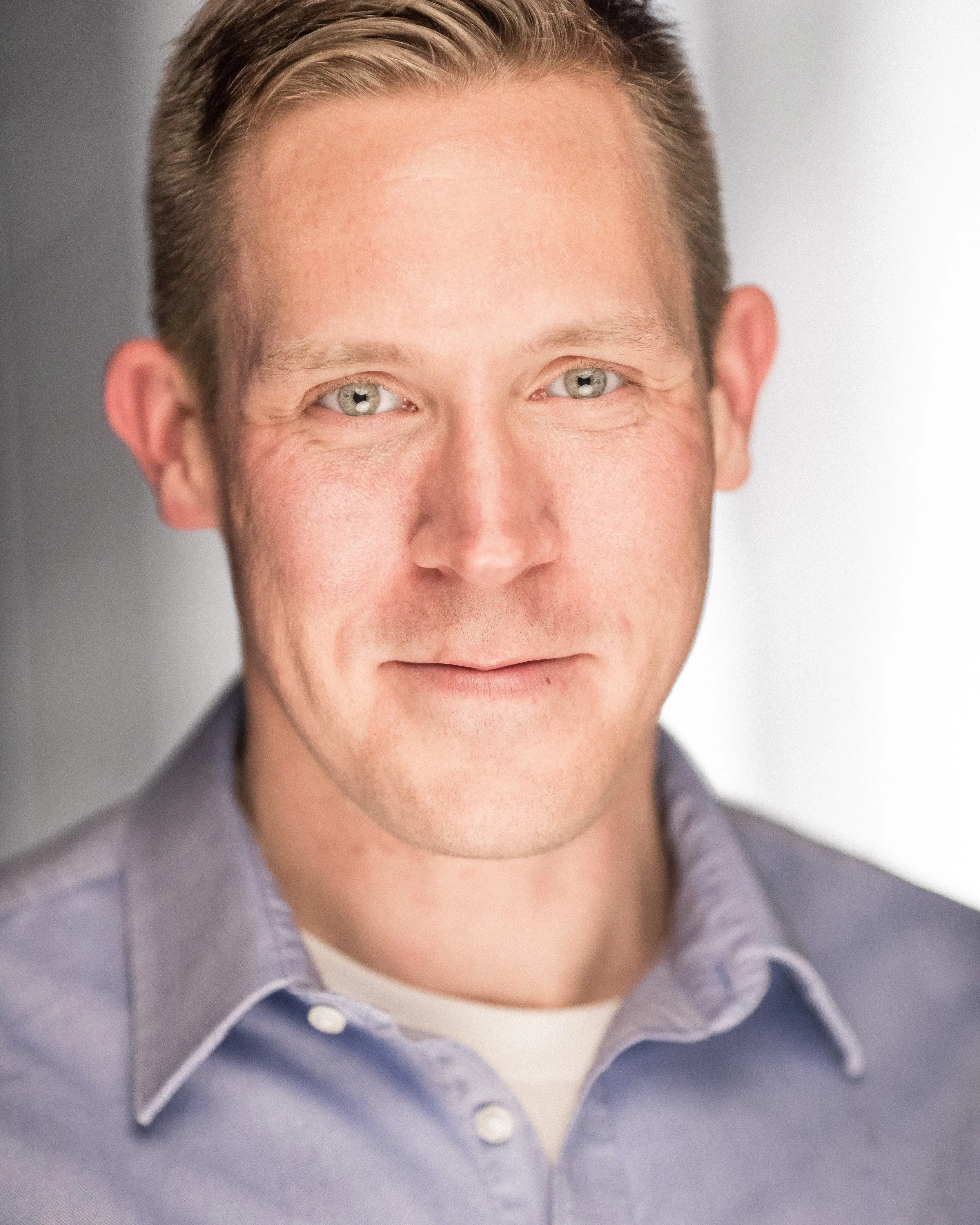 Close-up of a man's face with short blonde hair and blue eyes, wearing a light gray collared shirt over a white t-shirt.