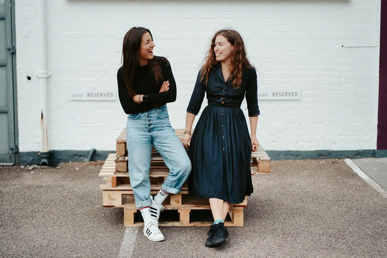 Two young women sitting on a wooden pallet in an outdoor urban setting, smiling and talking to each other.