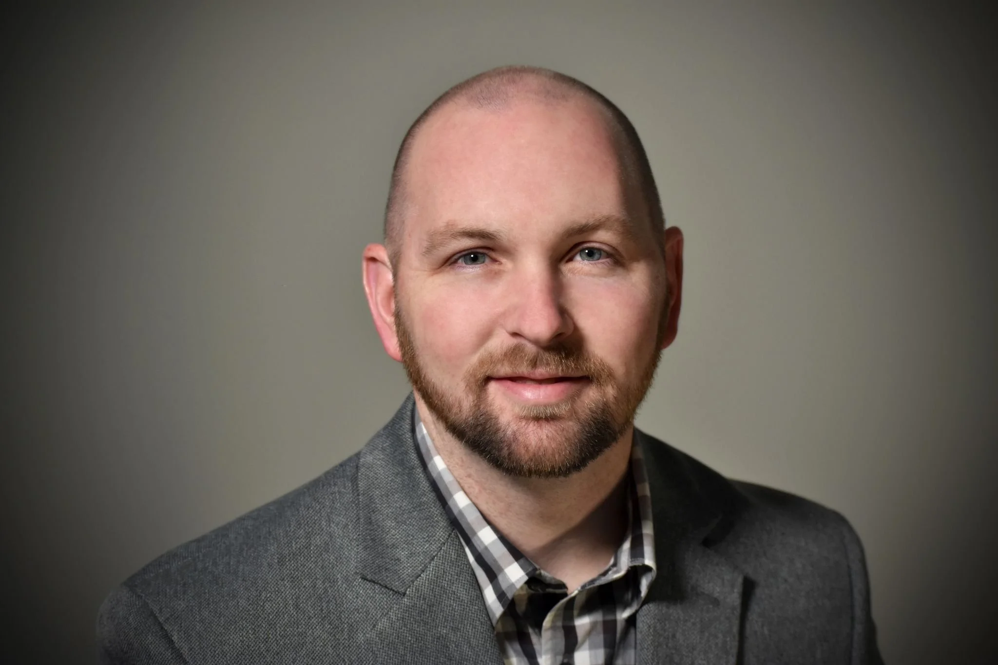 Close-up portrait of a man with a shaved head, light blue eyes, and a short beard, wearing a gray blazer over a checkered shirt, against a plain gray background.