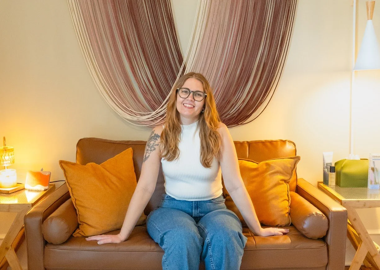 A woman with long red hair, glasses, and tattoos on her left arm smiling while sitting on a brown leather sofa with orange pillows in a warmly lit room. Behind her is a large, abstract wall hanging with flowing lines.