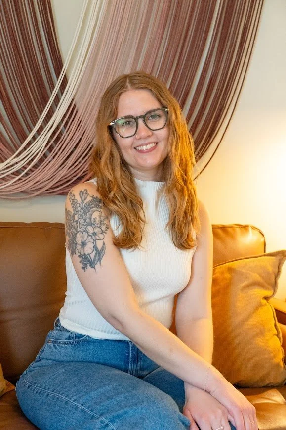 A young woman with long red hair, glasses, and a tattoo on her left arm, sitting on a brown sofa in a room with draped curtains behind her, smiling at the camera.