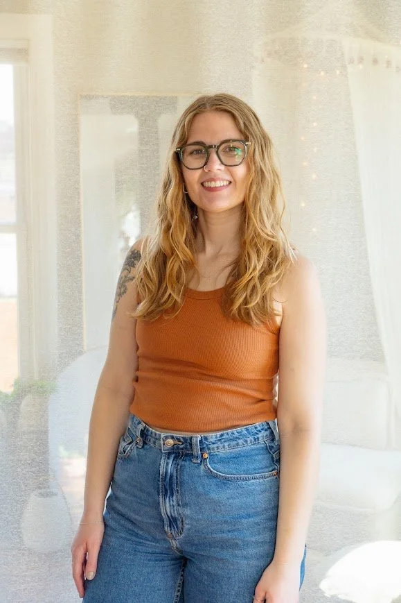 A young woman with long wavy blond hair, wearing glasses, a sleeveless orange top, and high-waisted blue jeans, standing indoors near a window with sunlight and string lights in the background, smiling at the camera.