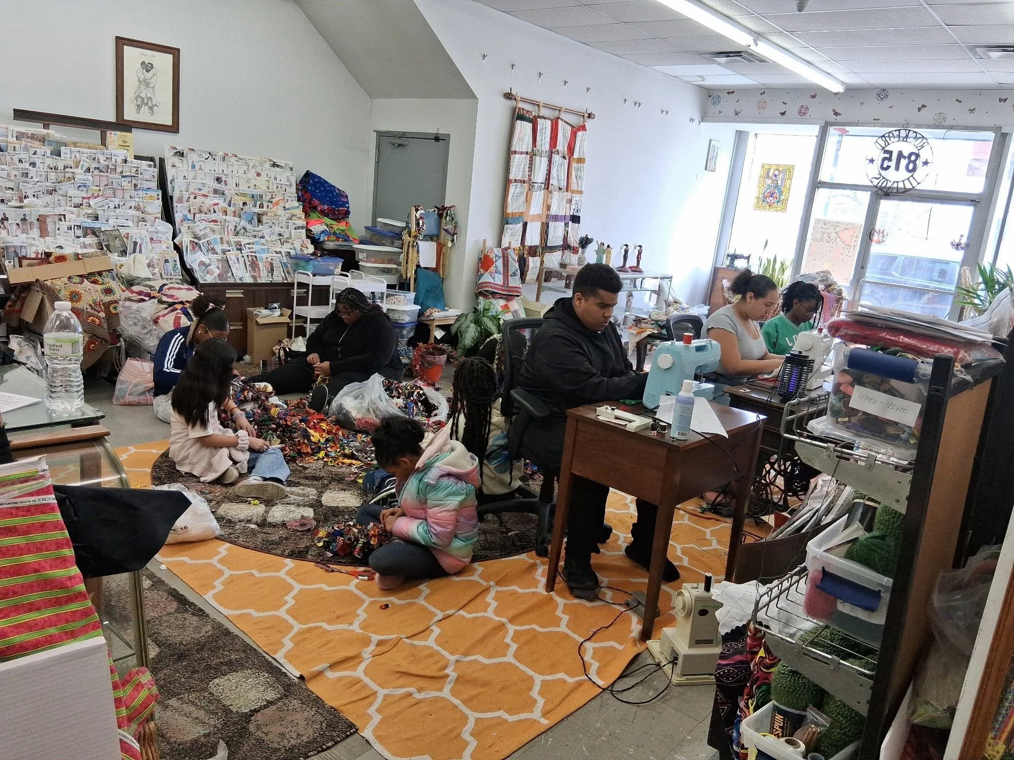 People organizing and sewing fabrics and textiles in a room, possibly for crafting or quilting, with shelves and tables filled with materials, a window letting in natural light, and a patterned orange and white rug on the floor.
