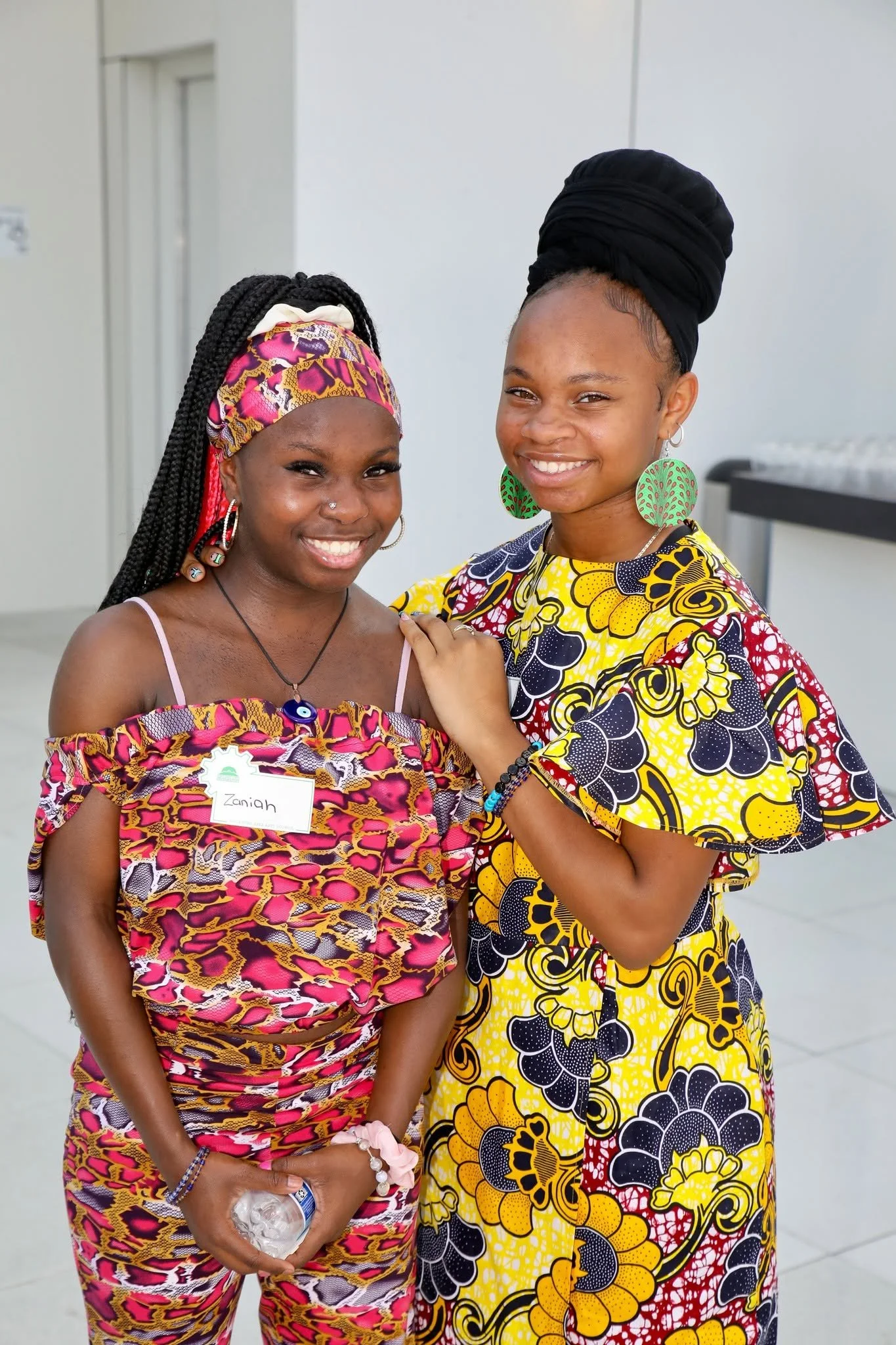 Two women dressed in vibrant, patterned clothing, smiling and standing close together indoors.