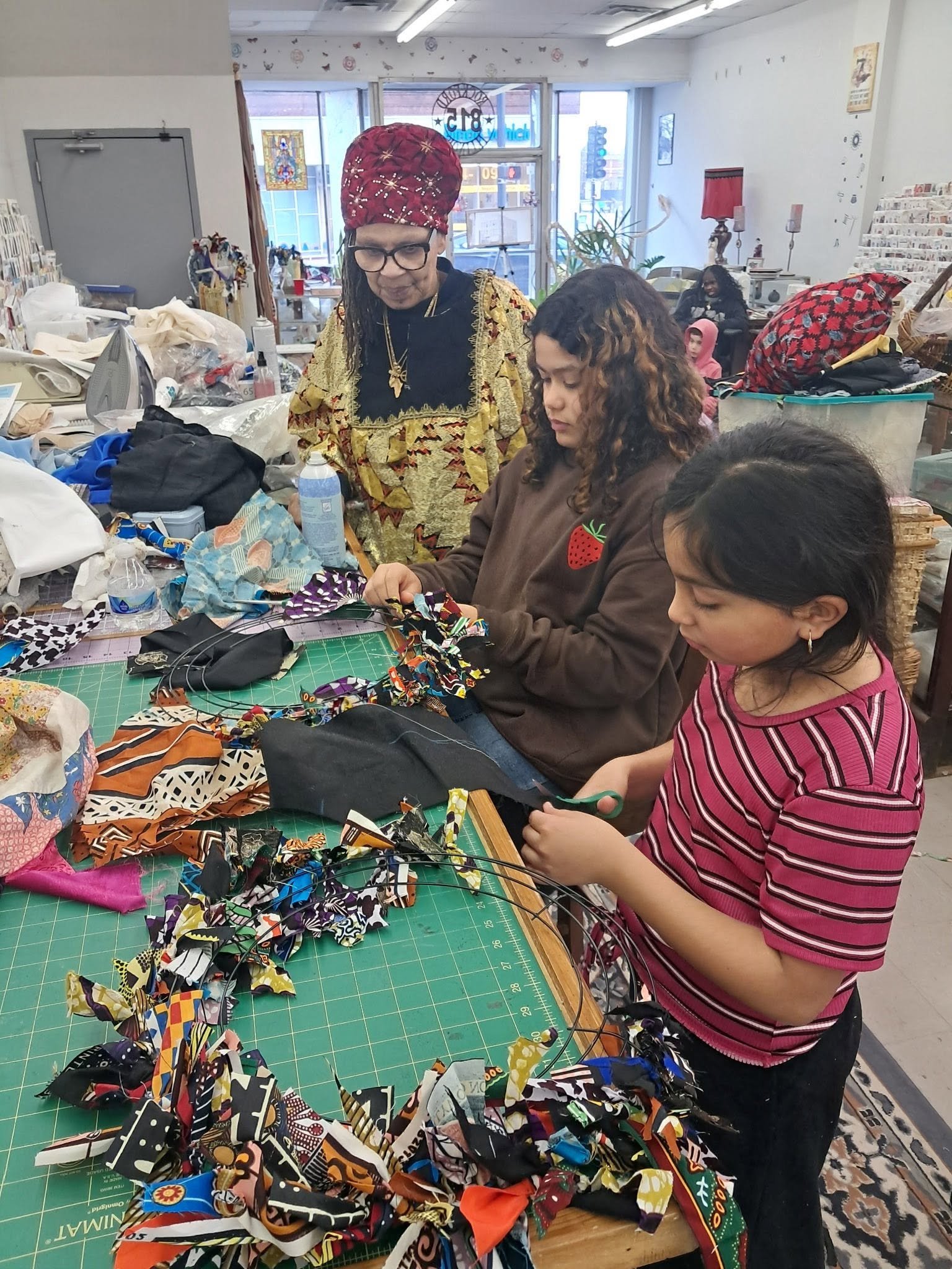 Three women working at a fabric crafting table filled with colorful fabric scraps and wires inside a bright store or workshop with shelves and curtains, and a person sitting in the background.
