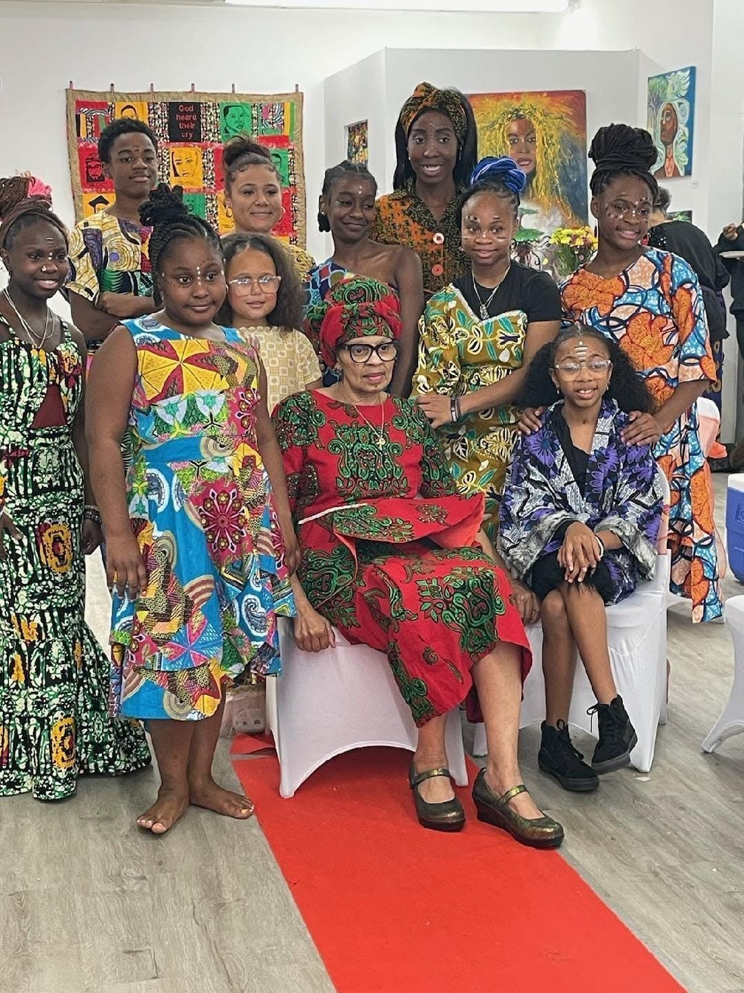 A group of diverse women and girls dressed in colorful African attire gathering at an art exhibit or cultural event. The woman seated in the center is wearing a red and green traditional dress with glasses, while the others stand around her, smiling.