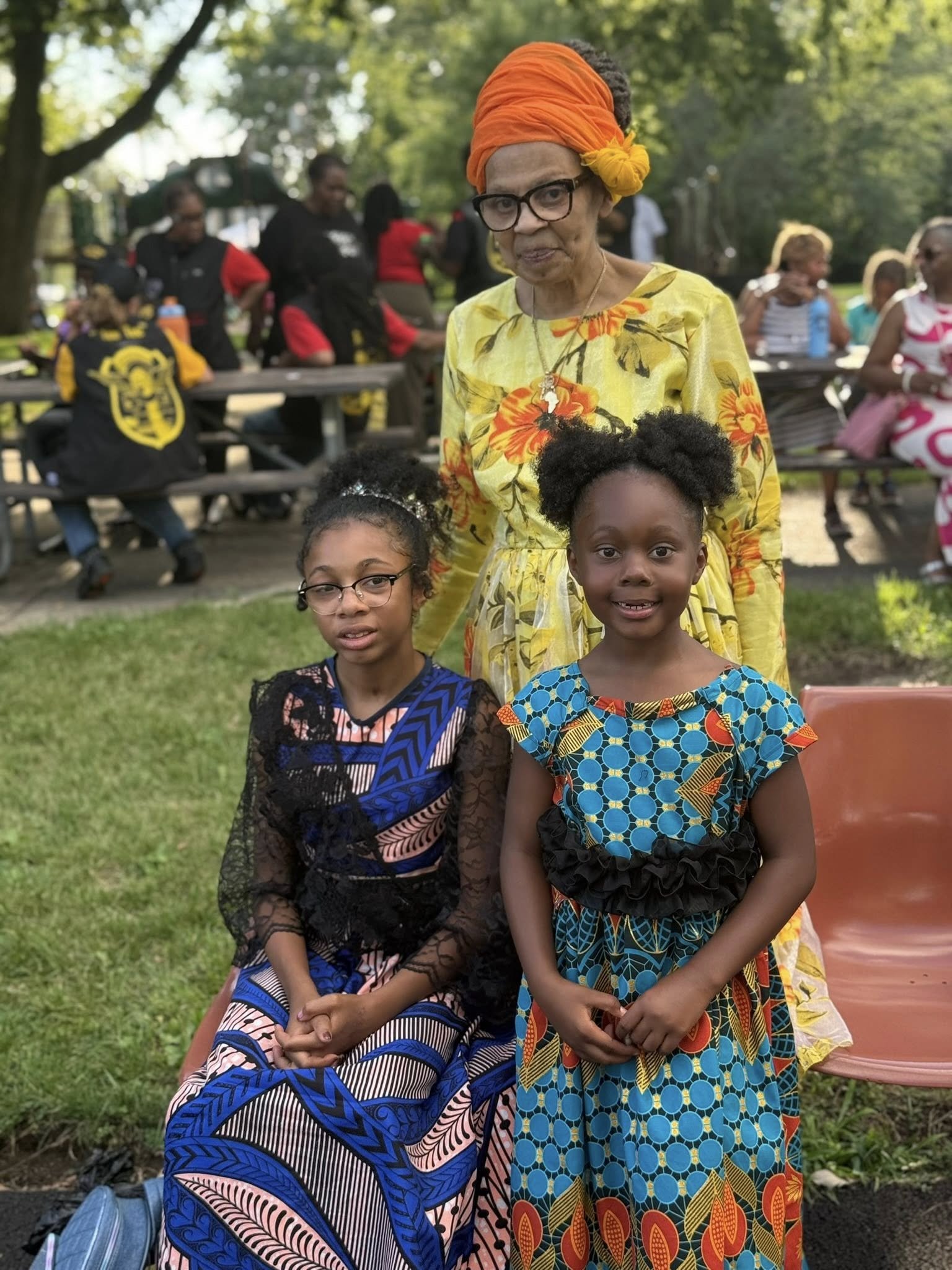 Three women and two young girls outdoors in a park, dressed in colorful African attire, with trees and other people in the background.