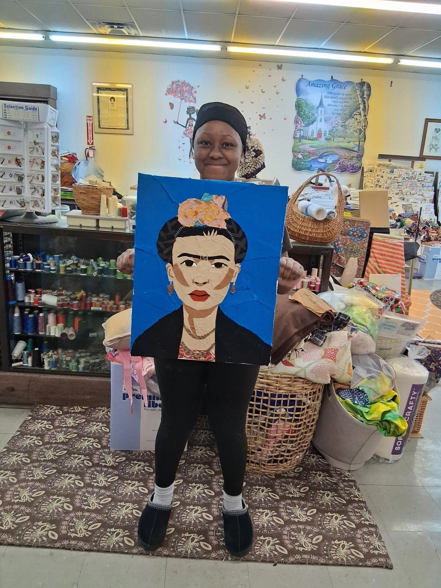 A person holding a quilting project featuring Frida Kahlo's face, standing in a craft store surrounded by sewing supplies and decorations.