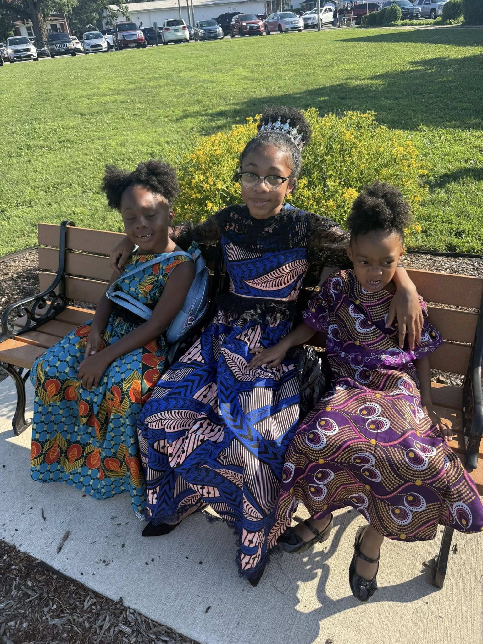 Three young girls sitting on a park bench on a sunny day, wearing colorful African dress, with a city street and parked cars in the background.