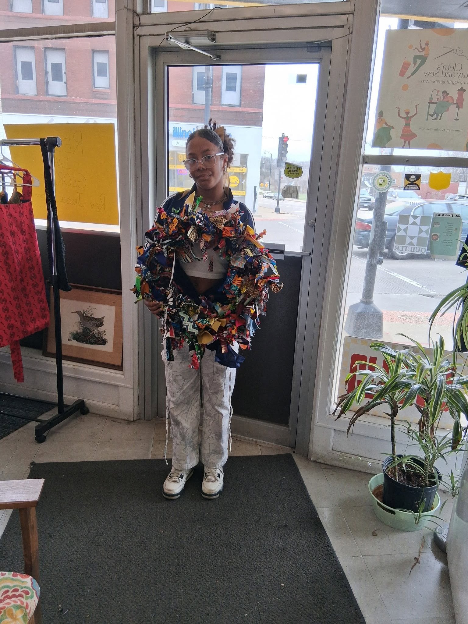 A person standing inside a store near a glass door, wearing a jacket with a colorful fabric pattern, white pants, and sneakers. The person has glasses and earrings. Outside the store, a street with cars, a traffic light, and buildings is visible thro