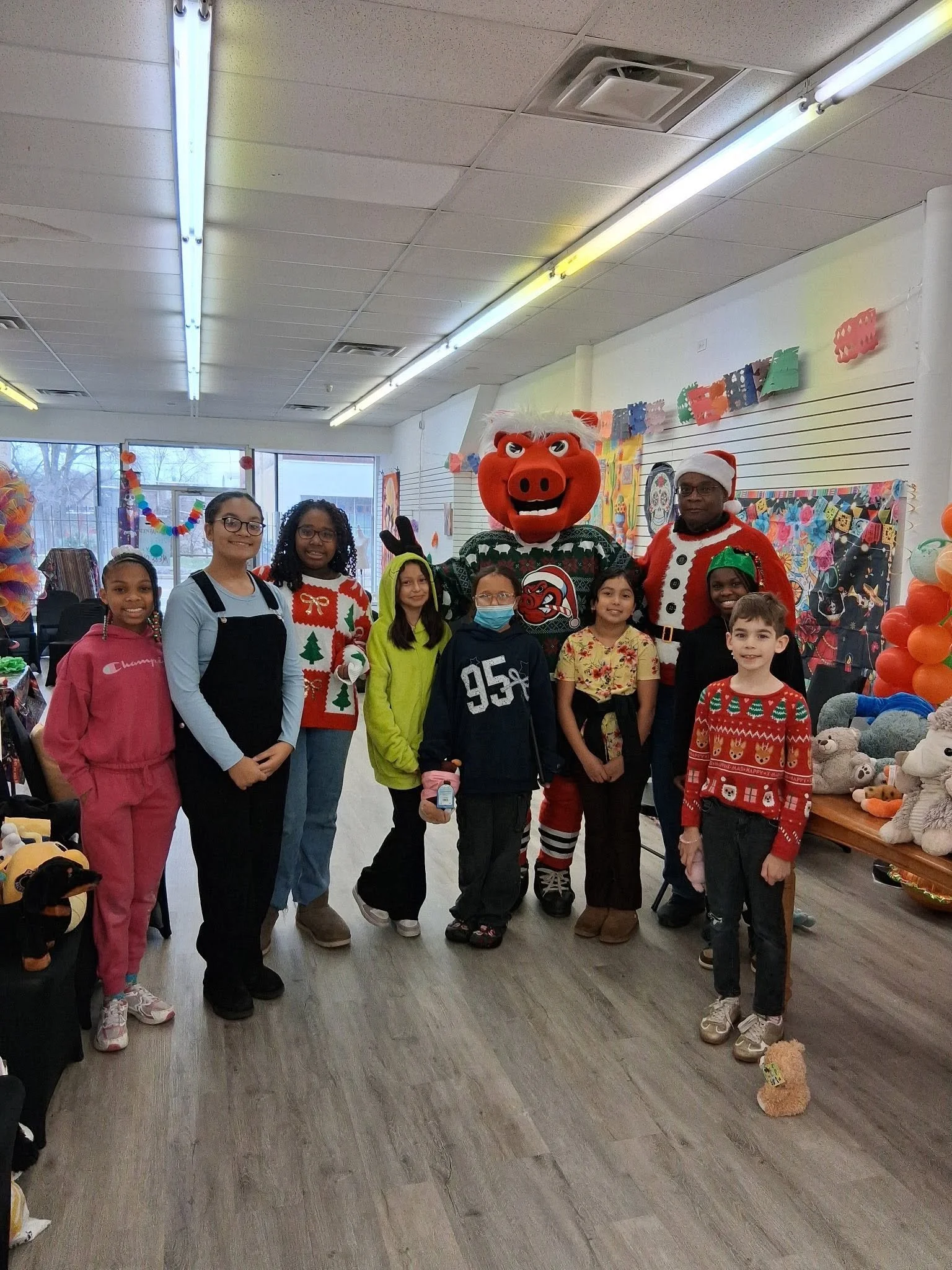 A group of children and two adults dressed in Christmas-themed costumes, including Santa hats and sweaters, standing indoors in front of a colorful decorations background, with a large pig mascot in the center.