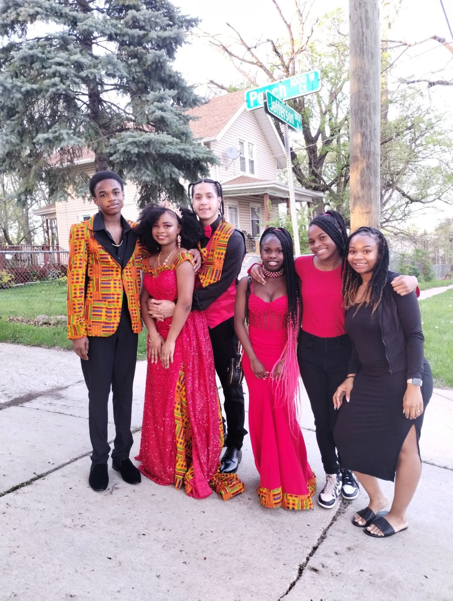 A group of seven young people standing together on a sidewalk in front of a house, with trees and street signs behind them. They are dressed in vibrant, colorful clothing and smiling at the camera.