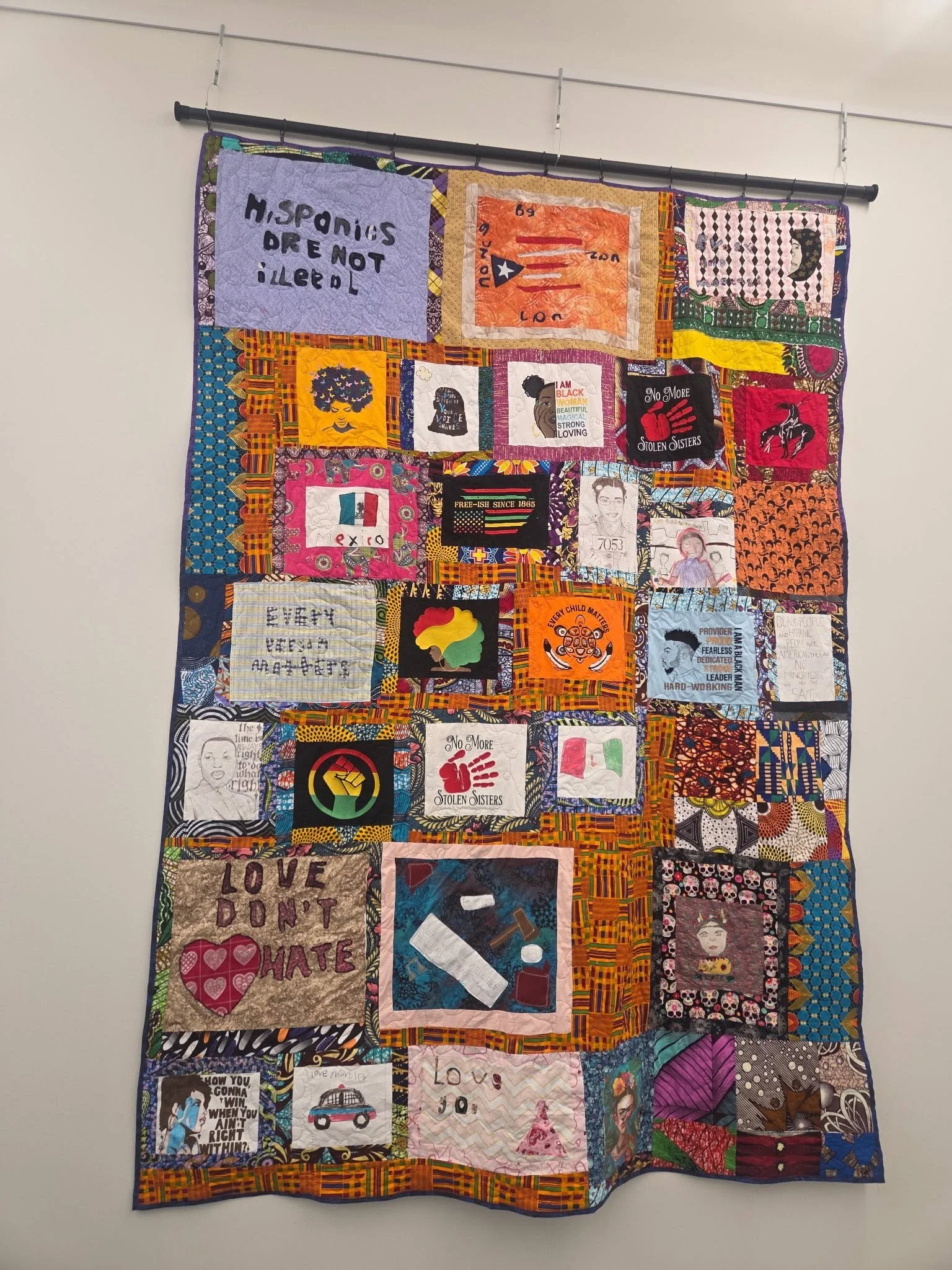 A colorful quilt hanging on a wall with various patches, including political messages, artwork, and symbols. Some patches feature slogans like "Hispanics are not illegal," "Love don't hate," and images of people, flags, and abstract designs.