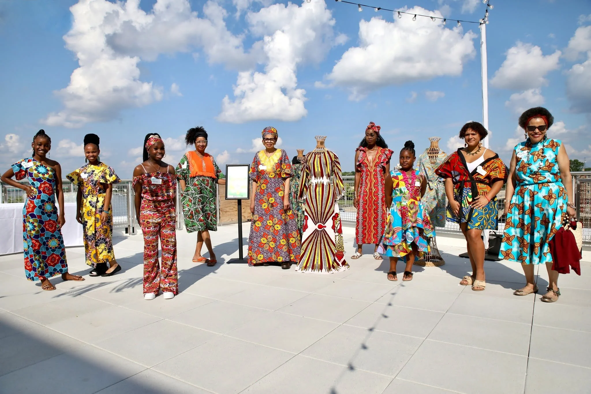 Group of women and girls dressed in colorful traditional African clothing, standing outdoors on a rooftop under a partly cloudy sky, with a string of lights overhead.