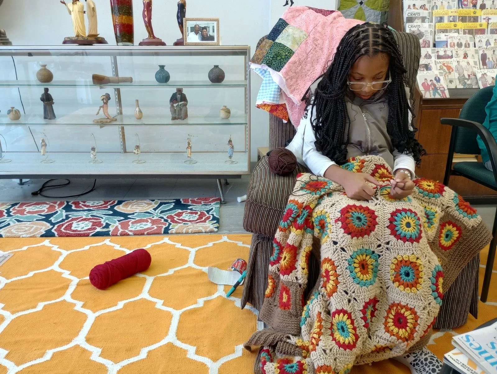A girl with glasses sitting in a chair crocheting a blanket with a colorful flower pattern. A ball of red yarn is on the yellow patterned rug in front of her, and she is surrounded by various craft supplies. Behind her, there is a display case with s