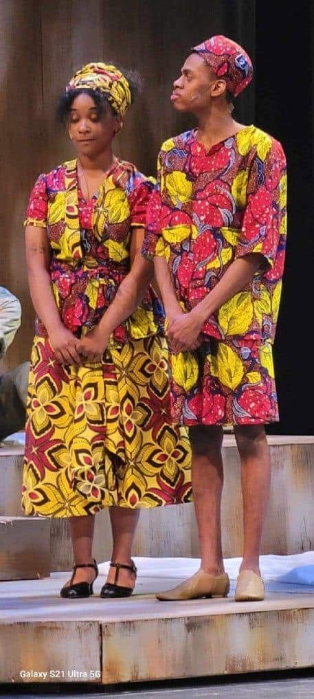 Two young women standing on stage wearing brightly colored, patterned African-style dresses and matching head wraps.