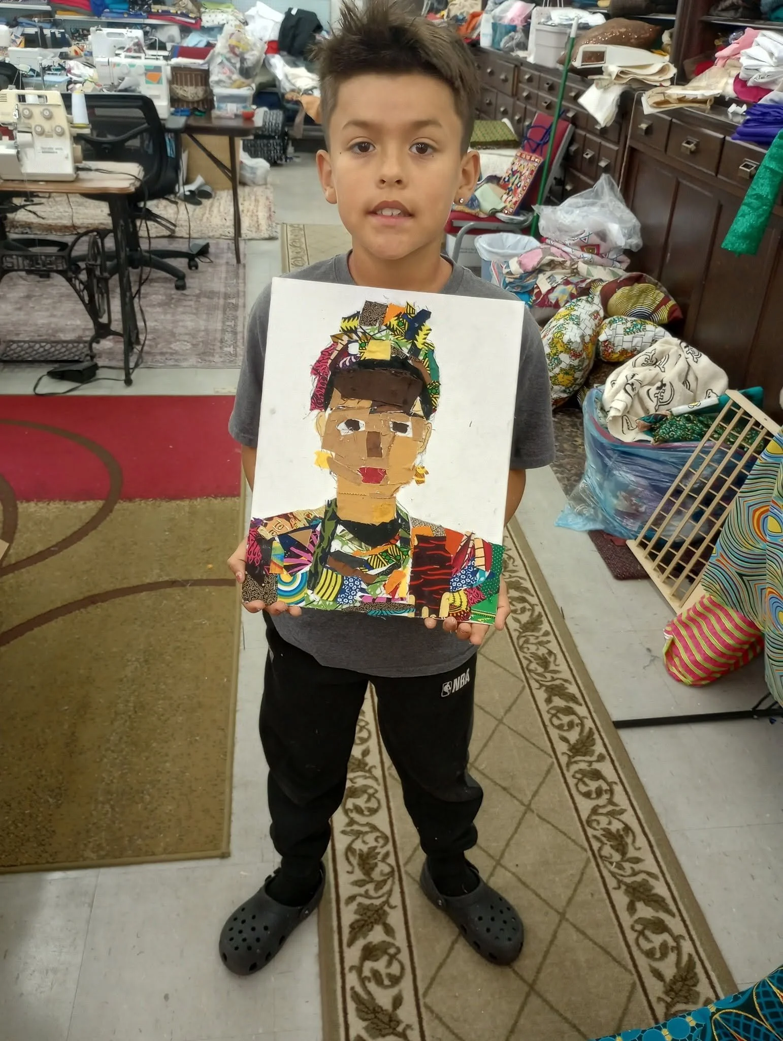 A young boy standing indoors in a cluttered room holding a colorful collage portrait of himself. The room has tables and shelves piled with fabric, books, and other items, with a sewing machine nearby.