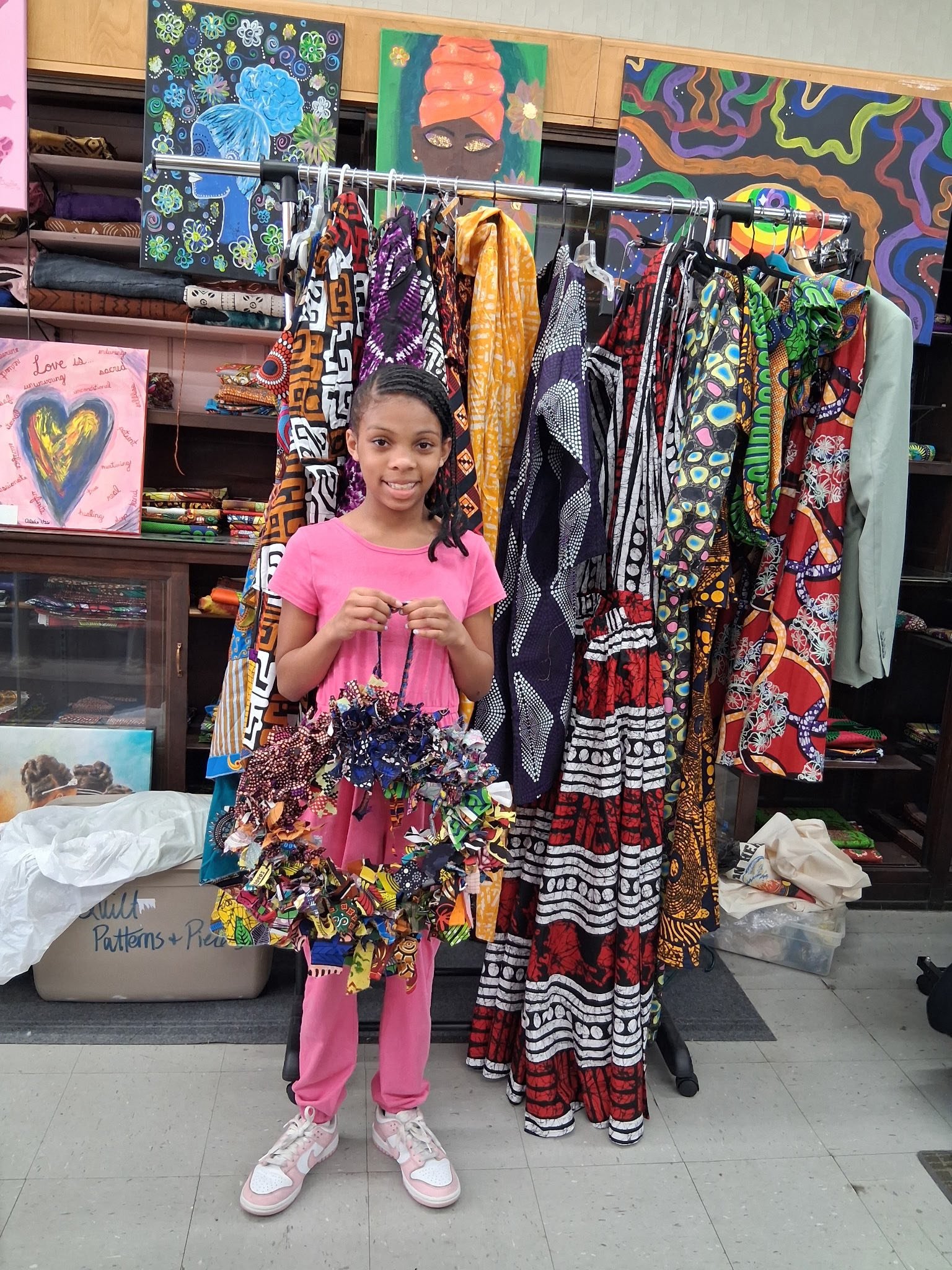 A young girl in pink pajamas and sneakers standing in front of a display of colorful clothing and artwork, including dresses with African-inspired patterns, in an indoor setting.