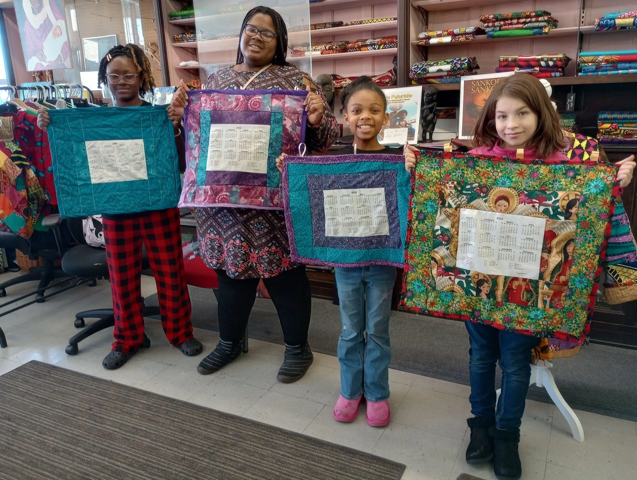 Four girls standing in a store, holding colorful fabric wall hangings with calendar prints in the middle. One girl wears red and black checkered pajamas and Crocs, one wears glasses and patterned clothing, another is in jeans with pink boots, and the last girl is partially visible on the right. Shelves of fabric are behind them.