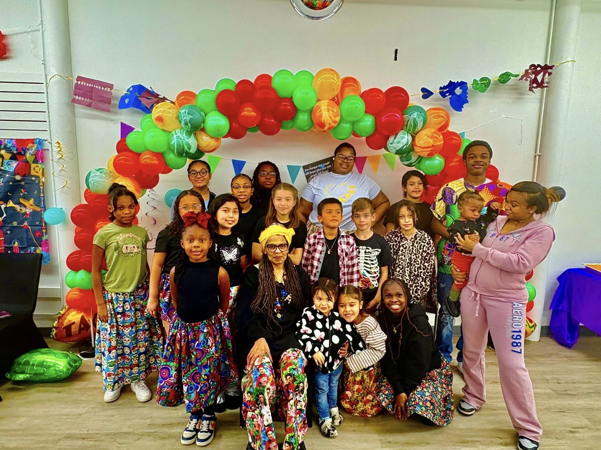 A group of children and adults smiling at a party decorated with colorful balloons and banners.