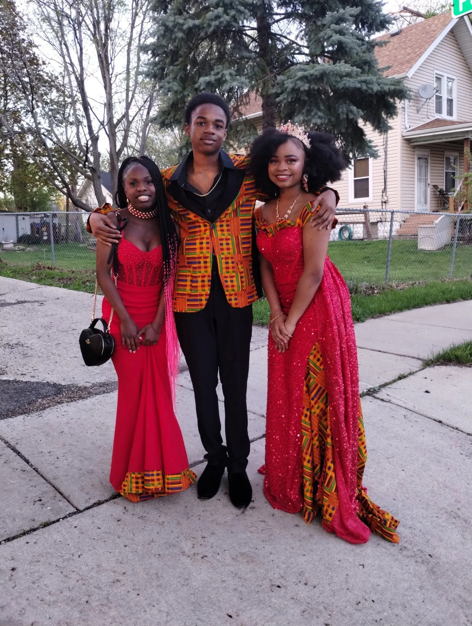 Three young people dressed in colorful African attire, standing on a sidewalk in front of a light-colored house with a fenced yard, during daytime.
