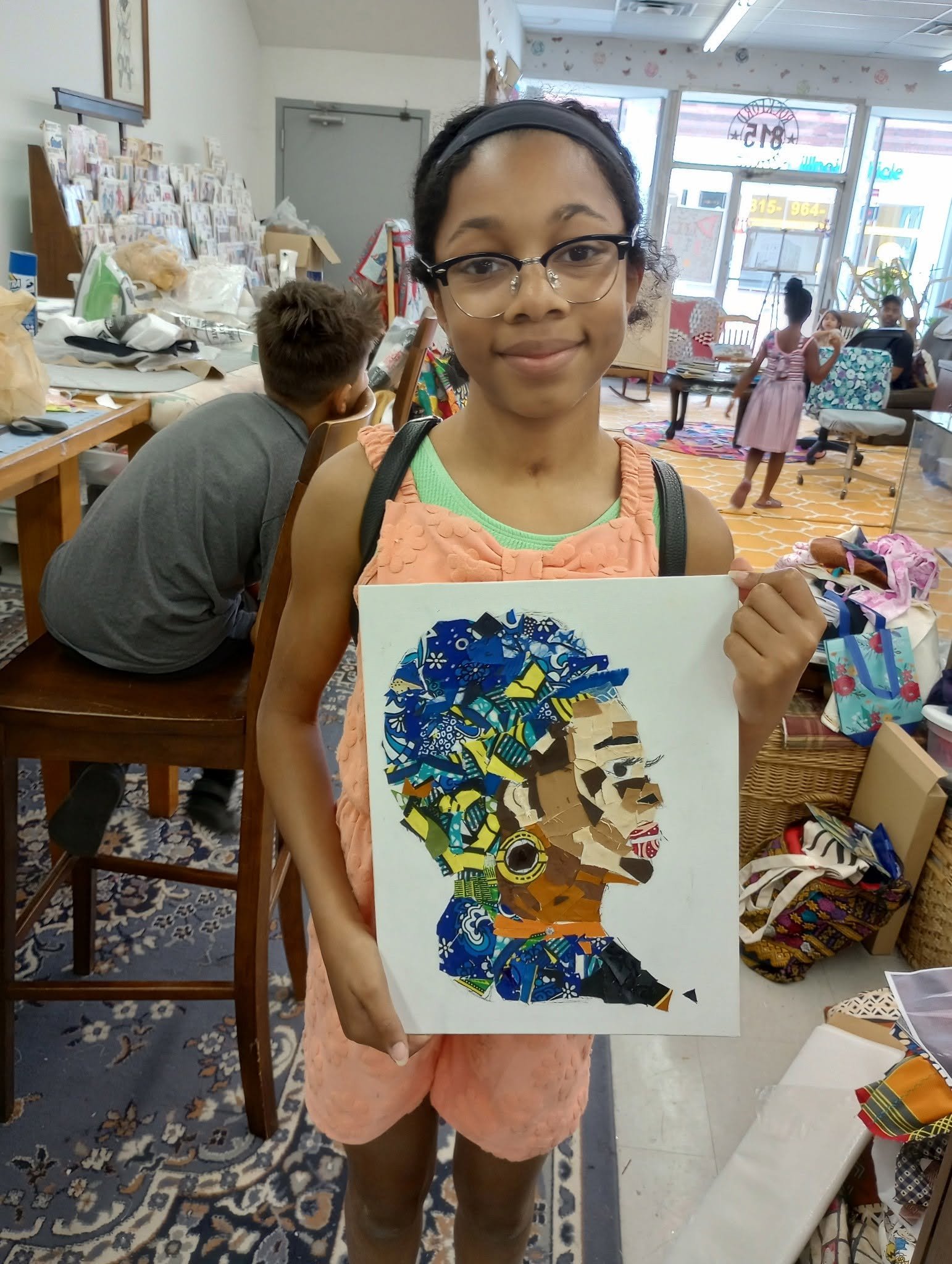 A young girl with glasses and a headband holding up an artwork made of colorful paper collage in an art studio. In the background, there are tables with art supplies, children, and a window showing the outside.