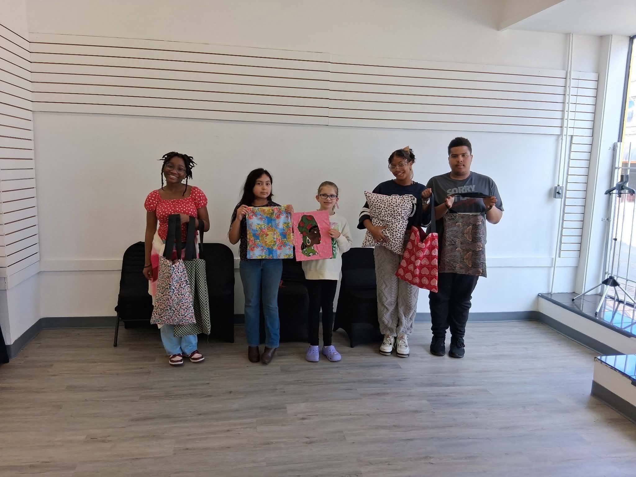 Six children standing in a row indoors, holding gift bags and artwork, smiling and facing the camera, with a white wall and window in the background.