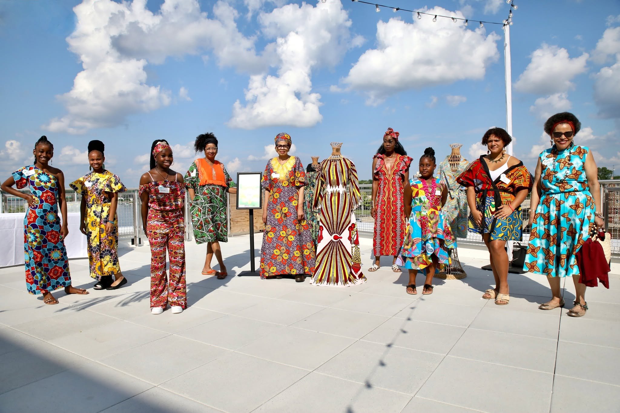 Group of diverse women dressed in colorful, patterned traditional African attire standing outdoors on a sunny day with a blue sky and clouds. There are decorative baskets and a framed display in the background.
