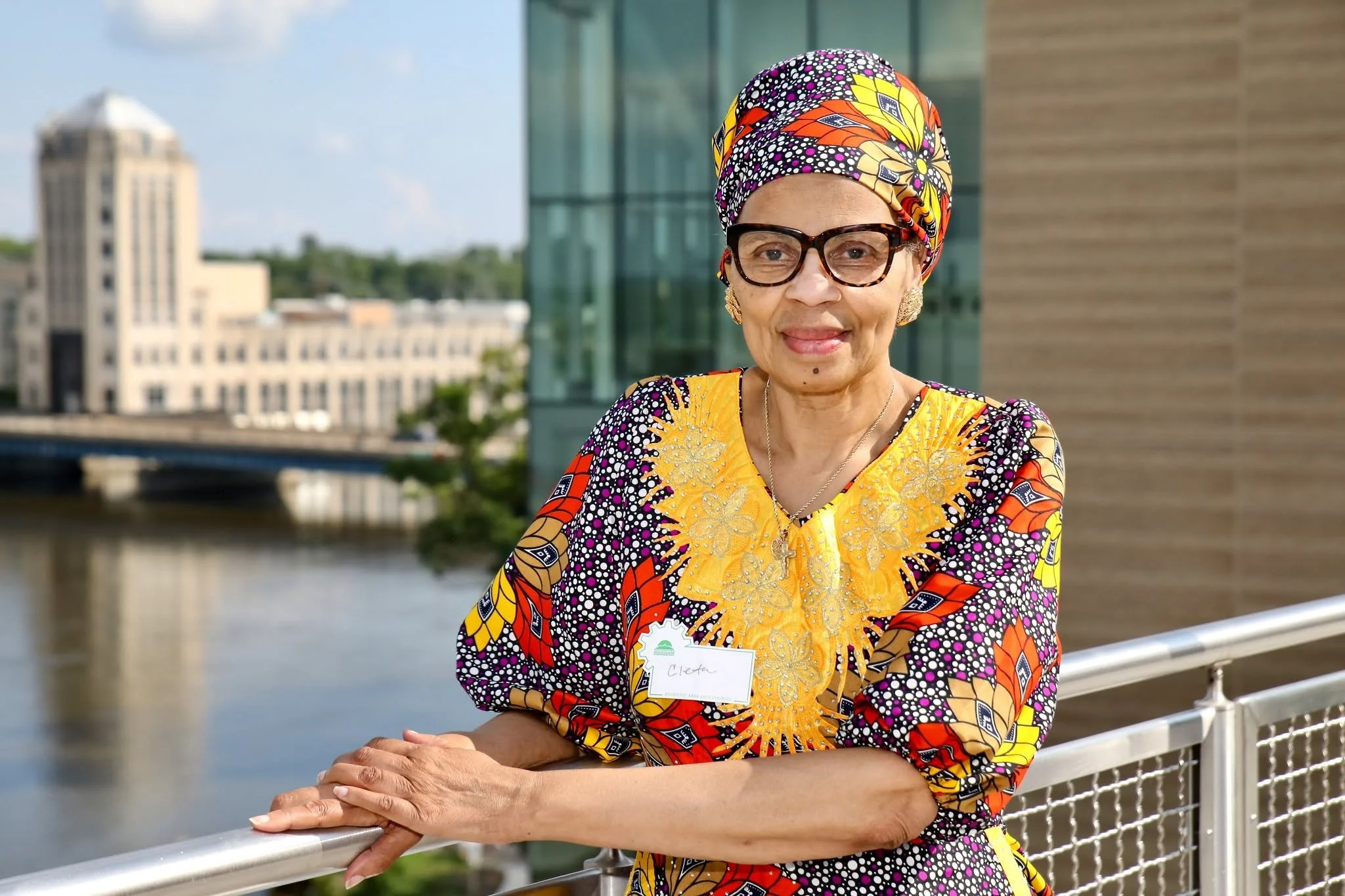 A woman wearing a colorful African print dress and matching headscarf, with glasses and earrings, posing on a balcony near a river with city buildings in the background on a sunny day.