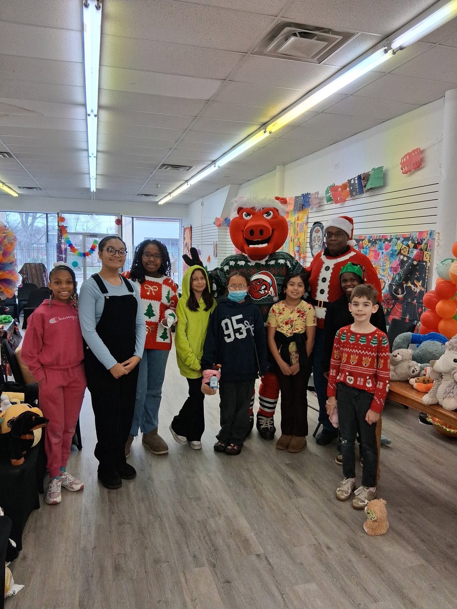 Group of children and two people dressed in Christmas costumes, including a pig mascot and Santa, in a decorated indoor space with toys and festive decorations.