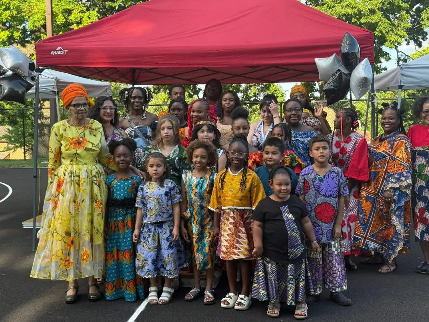 A group of children and adults dressed in colorful traditional African clothing pose together outdoors under a red canopy. The setting appears to be a park or playground with trees in the background.