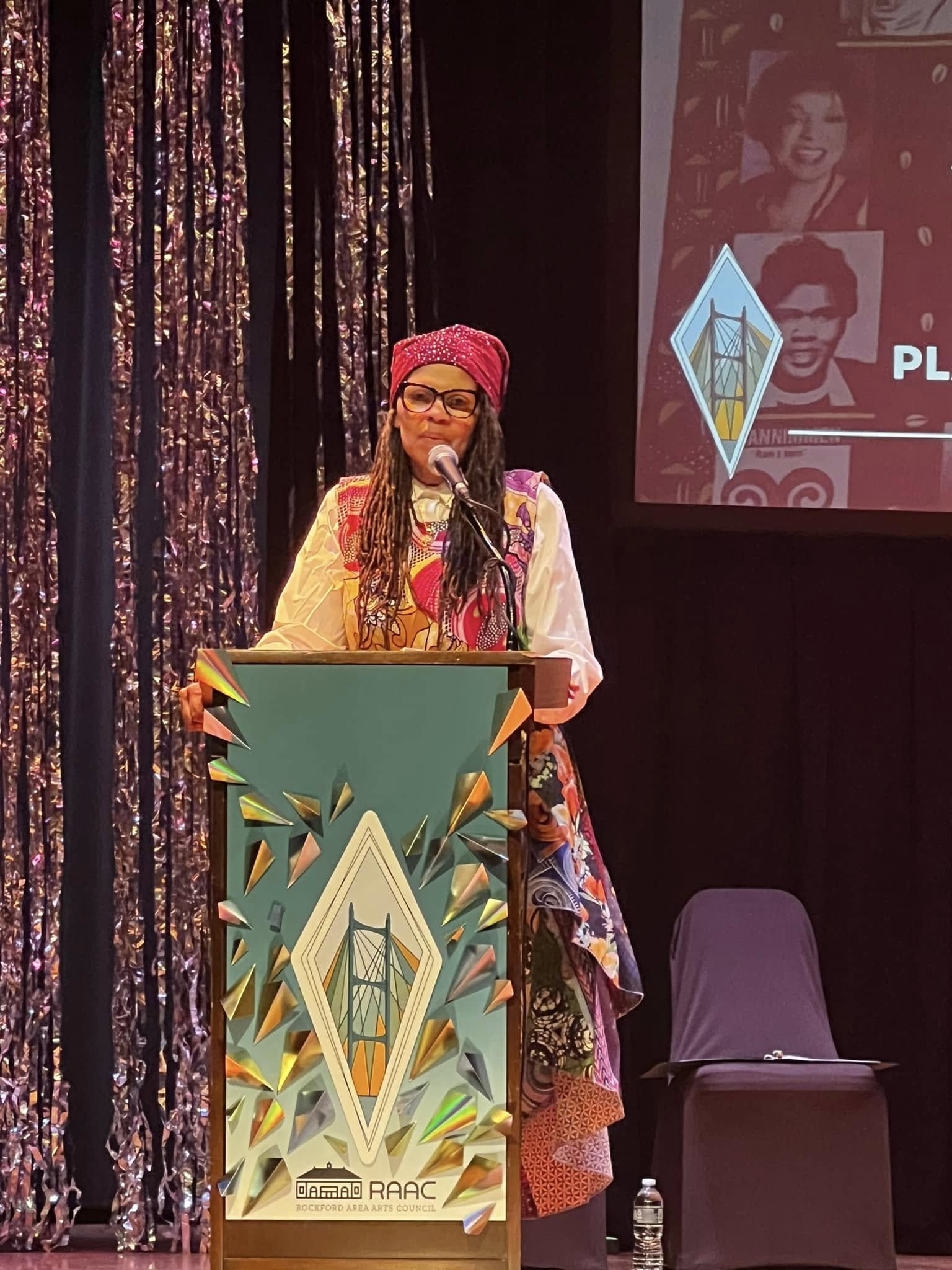 A woman with dreadlocks, glasses, and a bright pink headwrap stands at a podium, speaking into a microphone during an event. She wears a colorful patterned dress. Behind her, there is a large screen displaying a collage of photos and a logo for the R