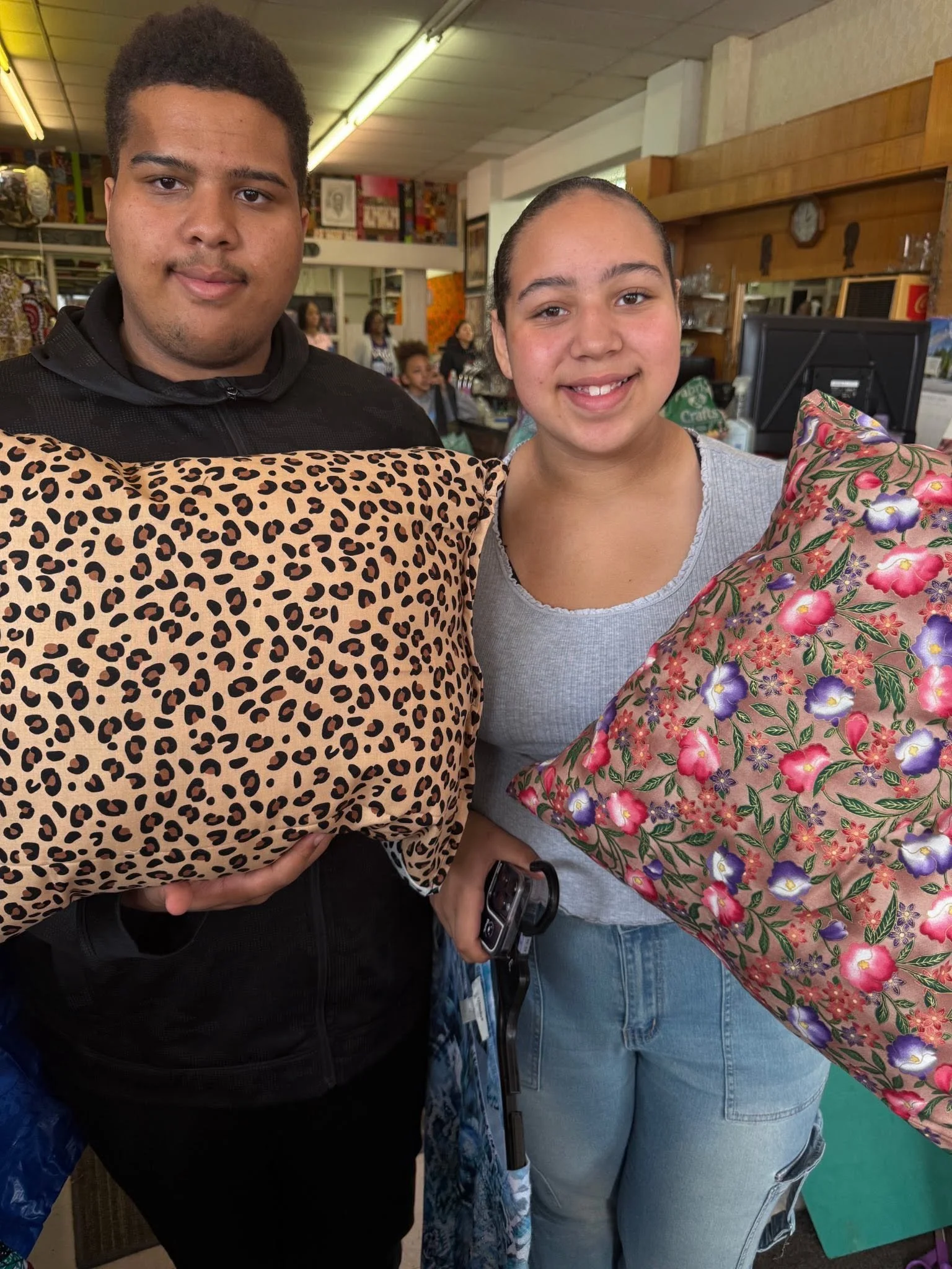 Two people standing in a store, holding colorful wrapped gifts. The person on the left is a young man with short dark hair, wearing a black top. The person on the right is a young woman with dark hair pulled back, wearing a gray t-shirt and light blue jeans. In the background, there are other shoppers, shelves, and store items.