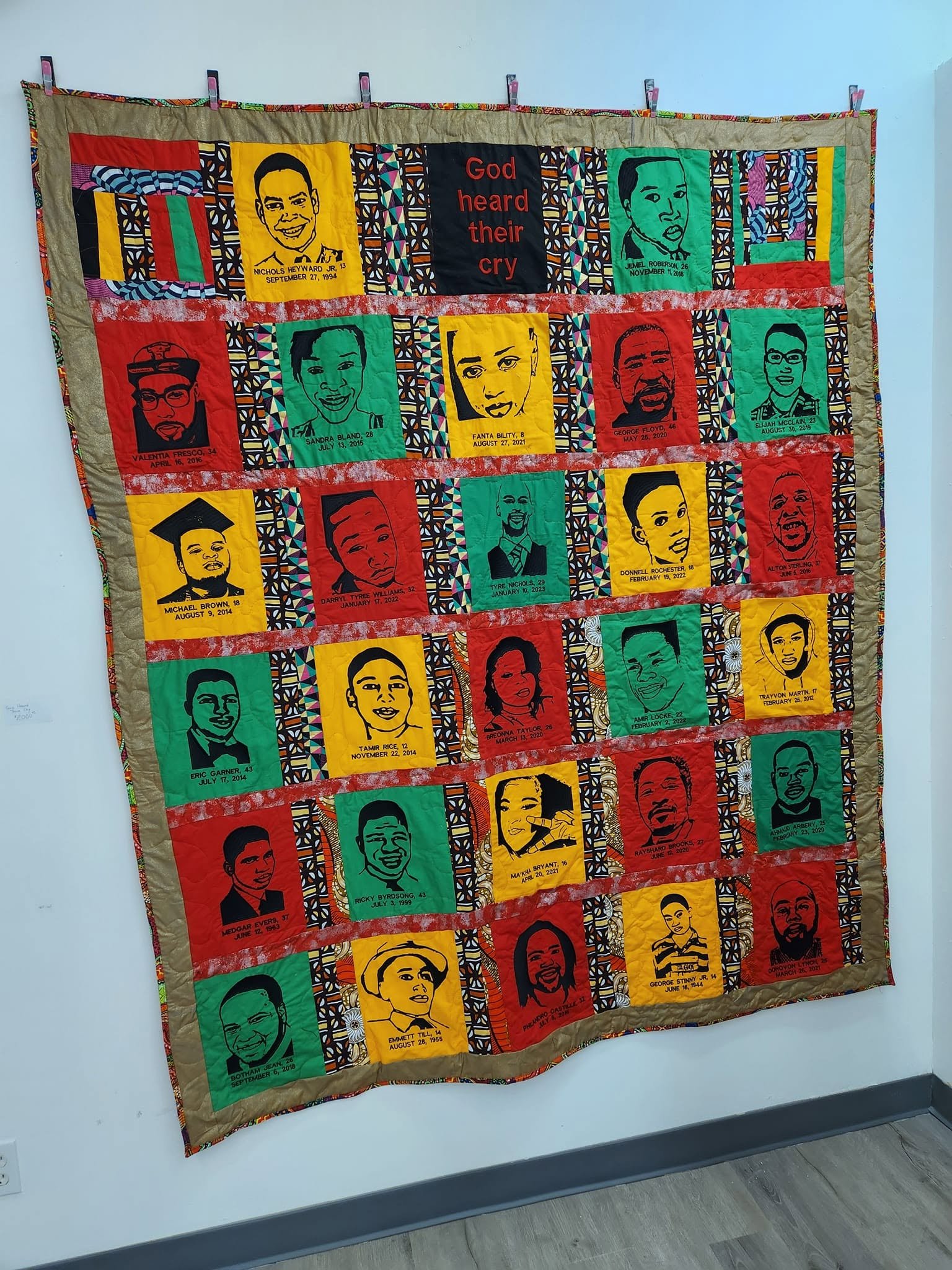 A quilt hanging on a wall featuring portraits of various individuals, with names and dates, and the phrase "God heard their cry" in the center.