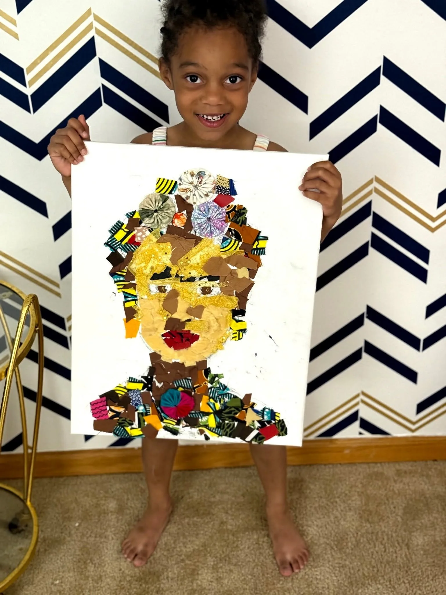 A young girl holding a colorful collage art piece of a woman's face in front of a geometric patterned wall.