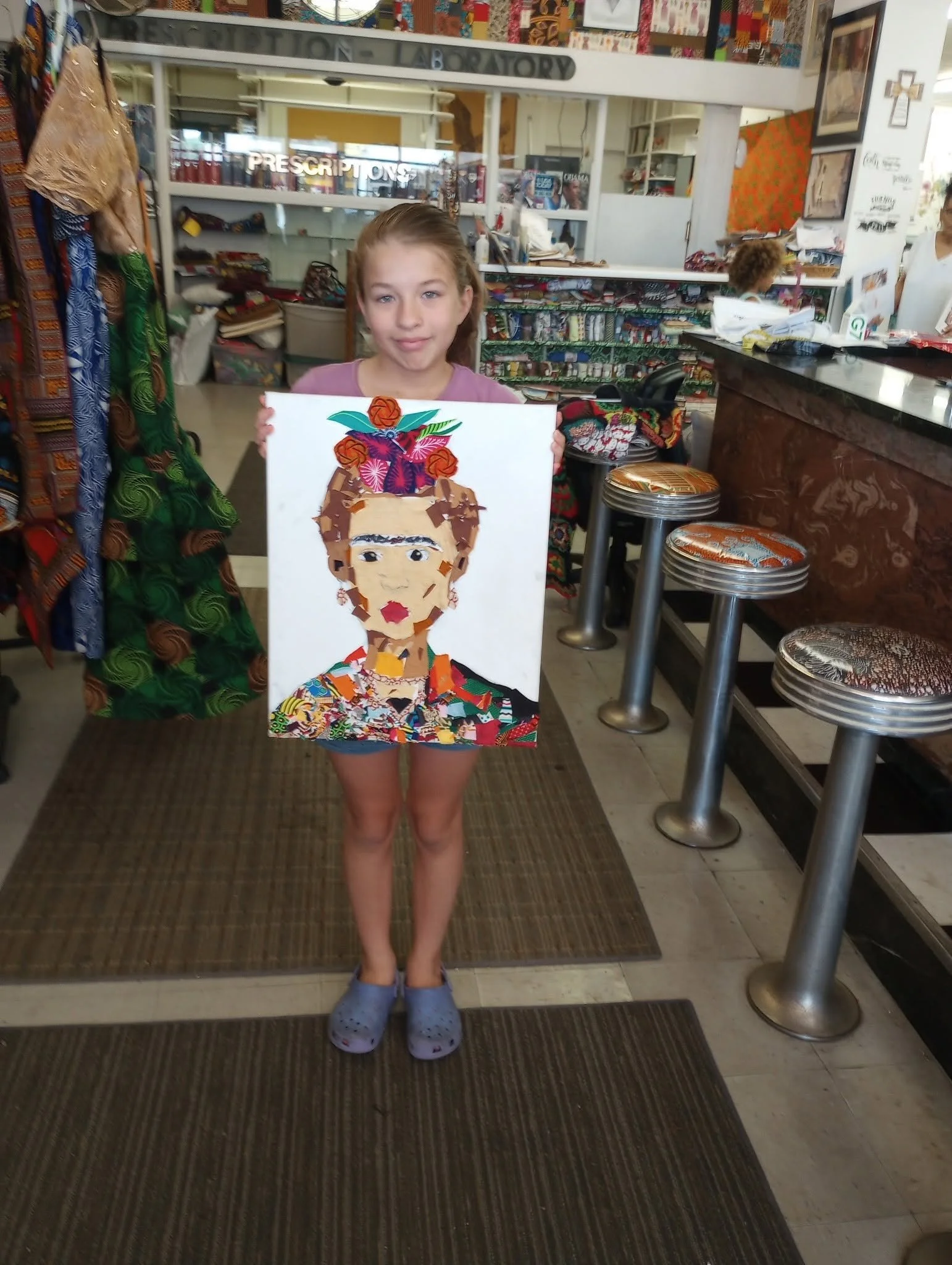 A young girl holding up a colorful collage art piece of a person's face inside a store or cafe.
