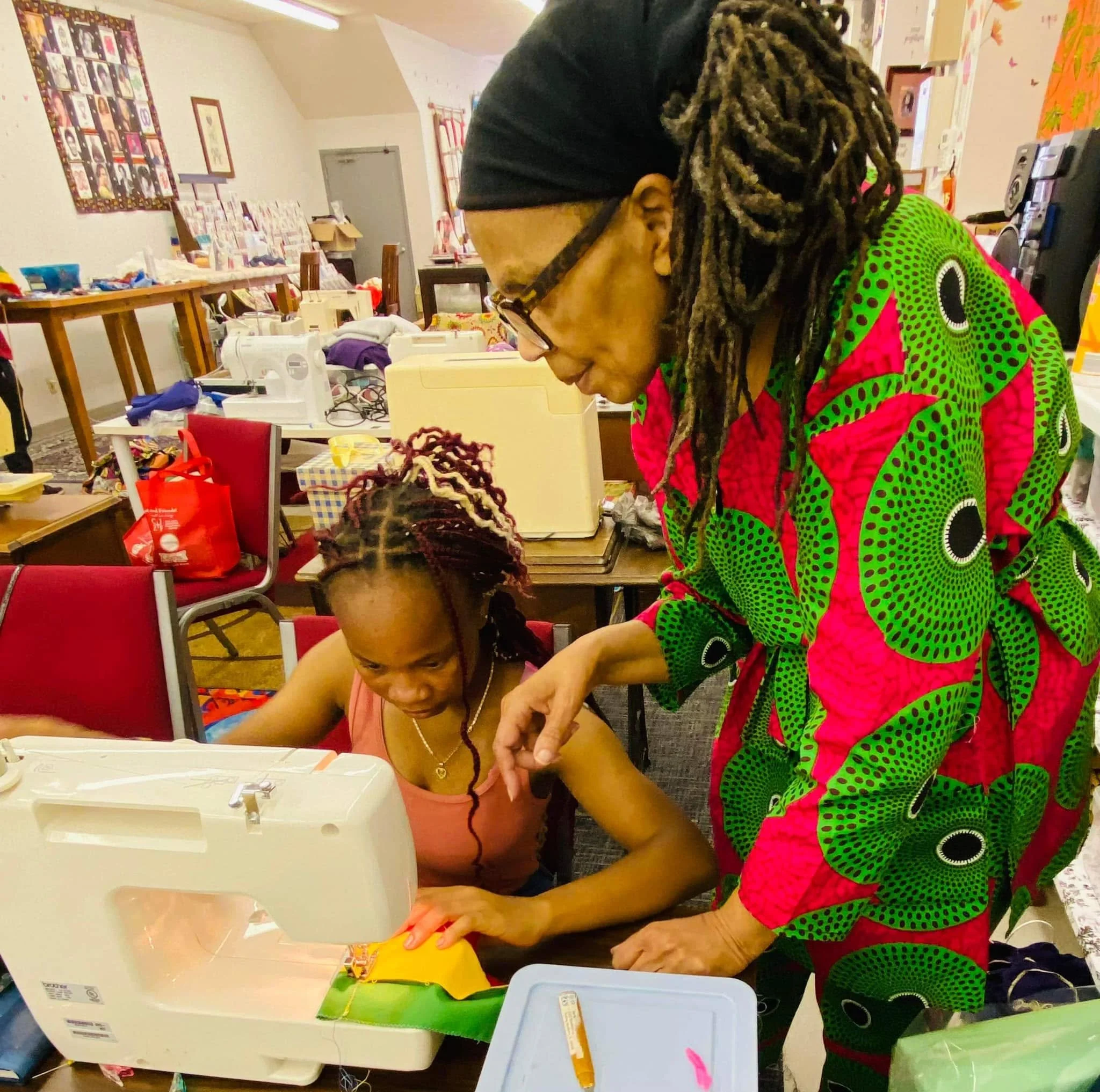 A woman with glasses and dreadlocks wearing colorful green and pink patterned clothing is sewing or crafting at a table with various supplies in a community room or classroom. Another person with a shaved head and a pink shirt is partially visible in